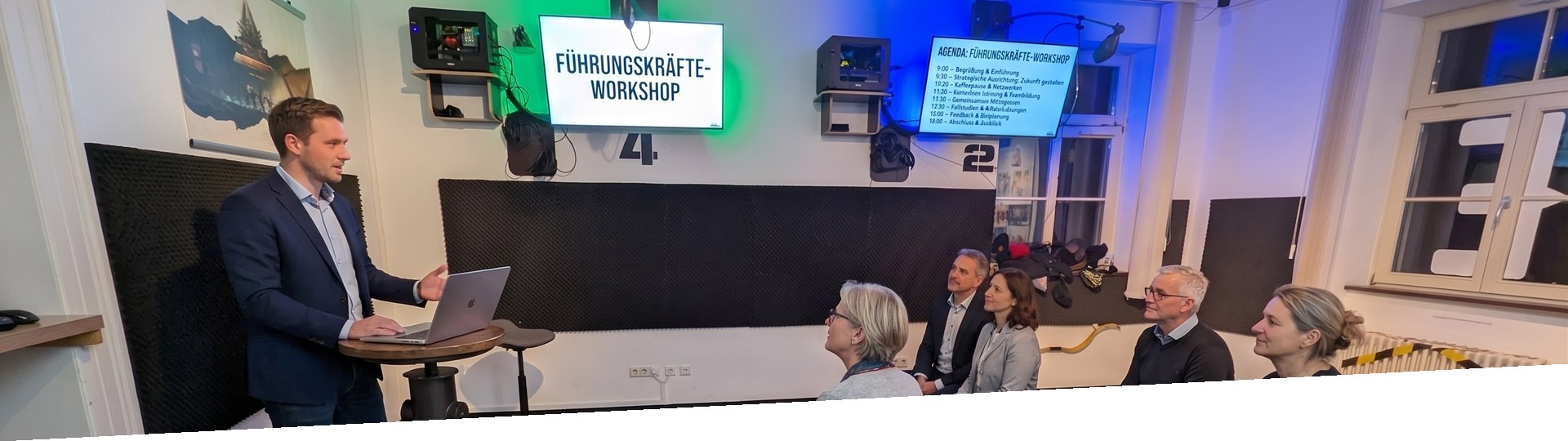 Fuehrungsteam im Workshop: Berater am Stehpult, Teilnehmende im Halbkreis, Bildschirm mit dem Titel Fuehrungskraefte-Workshop